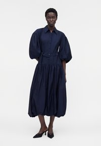 Femme portant une robe bleu marine ceinturée avec des manches bouffantes et un ourlet ballon froncé, associée à des escarpins noirs à bout pointu.