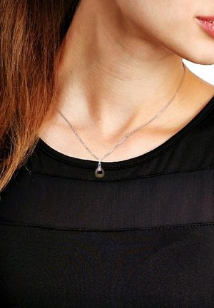 Collier en chaîne argentée avec un pendentif en forme de goutte noire, porté sur un haut noir, mettant en valeur un design simple et élégant.