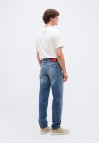 Νέος άνδρας όρθιος, κοιτάζει προς τα δεξιά, φοράει λευκό μπλουζάκι, μπλε τζιν με κόκκινο λογότυπο και μπεζ αθλητικά παπούτσια σε απλό φόντο.