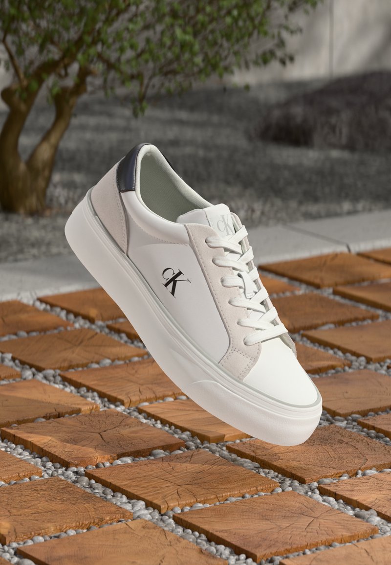 Witte leren sneaker met grijze suède accenten en een zwarte hiel. Beschikt over veters en een logo aan de zijkant. Vlakke rubberen zool.