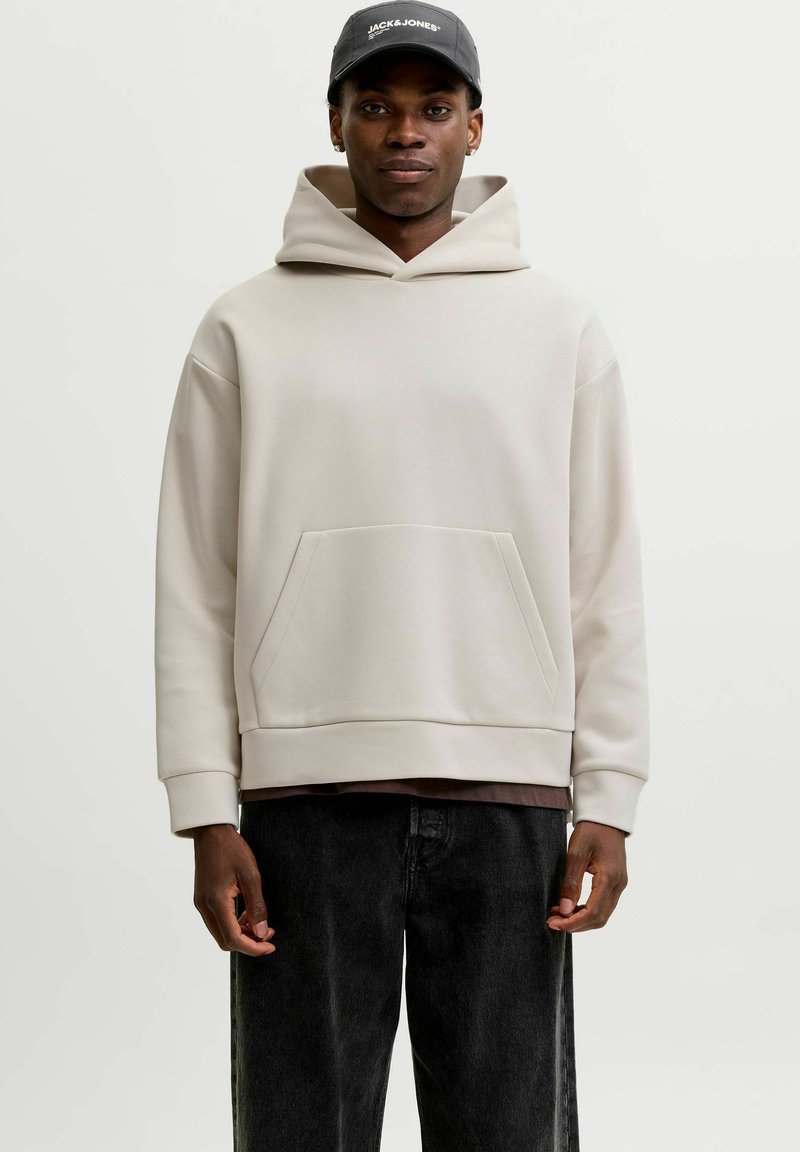Homme portant un sweat à capuche beige clair avec poche avant, jean noir et une casquette noire avec texte blanc, debout devant un fond clair uni.