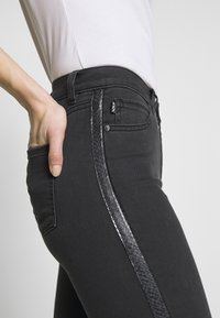 Los jeans skinny negros presentan un acento de estampado de serpiente texturizado a lo largo del costado, con un diseño clásico de cinco bolsillos y un acabado ligeramente desgastado.