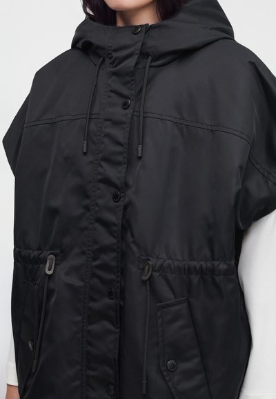 Veste noire à capuche avec boutons sur le devant, cordons ajustables à la capuche et à la taille, portée sur un haut blanc à manches longues.