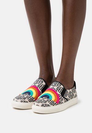 Nohy obuté v nazouvacích teniskách s vícebarevným duhovým vyšíváním a černým textovým vzorem na bílém pozadí.