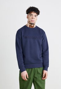 Hanorac bleu marin cu guler rotund și manșete ribbate, având un logo „Timberland” estompate pe piept.
