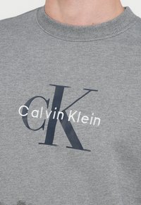 Šedá mikina s okrúhlym výstrihom, s výrazným tmavomodrým logom "CK" a bielym nápisom "Calvin Klein". Mäkká textúra, ležérny dizajn.