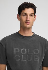 Camiseta corta de manga en gris oscuro, hecha de algodón y con un acabado texturizado. Presenta la frase "POLO CLUB" impresa en una fuente audaz y apagada en el frente.
