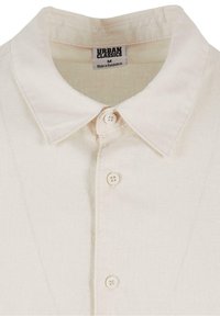 Chemise col crème avec trois boutons visibles, portant une étiquette Urban Classics indiquant la taille M, fabriquée au Bangladesh.