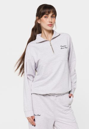 Kvinde med langt brunt hår iført en lys grå lynlåssweatshirt og matchende sweatpants med teksten "Sports Club" på begge.