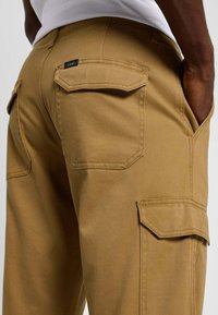 Pantaloni cargo beige realizzati in un tessuto liscio e testurizzato, con due grandi tasche applicate sui lati e un'etichetta sopra la tasca destra.