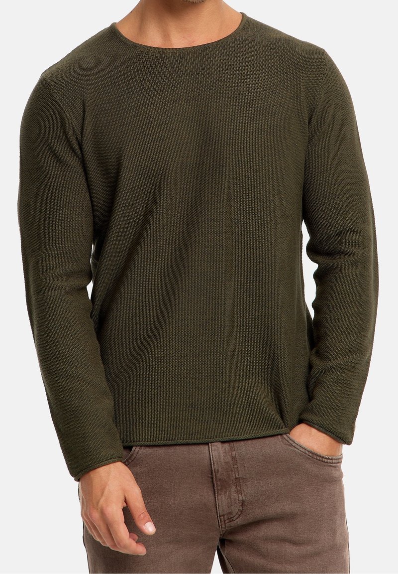 Homme portant un pull en tricot vert olive et un pantalon marron, une main dans la poche, avec un fond neutre.