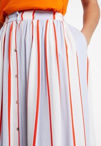 Pruhovaná midi sukně v světle modré a bílé barvě, zdobená výraznými červenými pruhy, s knoflíkovým zapínáním vpředu a bočními kapsami z lehké látky.