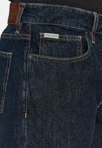 Tamno plavi traperice s teksturiranom površinom, s zlatnim šavovima i kožnom oznakom koja na pojasu ističe "GUESS JEANS".