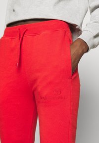 Red sweatpants feature a drawstring waistband, side pockets, and an embroidered logo. The fabric has a smooth texture and relaxed fit.