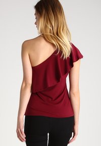 Top burdeos de un solo hombro con un acento de volantes; diseño ajustado con textura suave, combinado con pantalones negros.