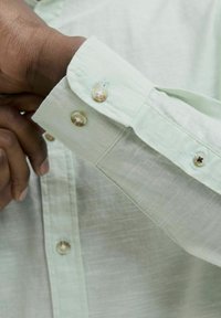 Manșeta unei cămăși lungi cu mânecă, de culoare verde deschis, texturată, cu două butoane rotunde, de culoare deschisă, având o închidere cu un singur buton și dintr-un material moale.
