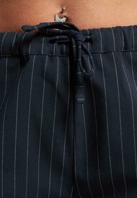 Marineblå bukser med pinstriper i stoffet og strikk i livet. Har sølvfarget utstyr og en liten sommerfugldetalj på livet.