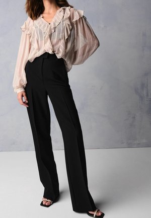 Blouse légère rose pâle, transparente, avec des détails volantés ; associée à un pantalon noir taille haute, ajusté ; texture lisse et coupe ample. Sandales noires.