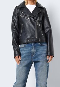 Kobieta w czarnej skórzanej kurtce motocyklowej z srebrnymi zamkami błyskawicznymi i niebieskich jeansach, stojąca na jednolitym jasnym tle.
