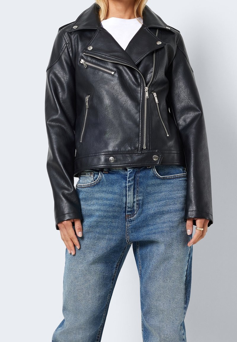 Kobieta w czarnej skórzanej kurtce motocyklowej z srebrnymi zamkami błyskawicznymi i niebieskich jeansach, stojąca na jednolitym jasnym tle.