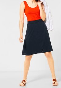 Červený top bez rukávů spárovaný s tmavě námořnickou sukní s překládaným designem, vystavený s hnědými sandály na vyšším podpatku na jednoduchém pozadí.