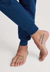 Blauwe sportbroek met elastische boorden, gecombineerd met blote voeten met meerdere tatoeages. De huidtextuur is zichtbaar.