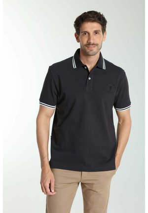 Homme portant un polo noir avec des rayures blanches sur le col et les manches, associé à un pantalon beige, debout sur un fond uni.