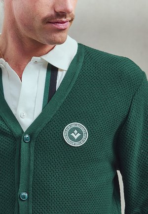 Groene netstructuur cardigan met een V-hals, voorzien van een rond logo-embleem, knoopsluiting en een gestructureerd patroon. Gelaagd over een witte polo-kraag.