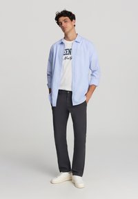 Jasnoniebieska koszula z długim rękawem na białym t-shircie z grafiką, w połączeniu z ciemnoszarymi spodniami i białymi trampkami. Gładki materiał z podwiniętymi rękawami.