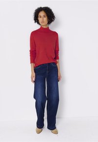 Kobieta stoi na białym tle, ubrana w czerwoną bluzę z golfem, ciemnoniebieskie dżinsy o szerokich nogawkach oraz beżowe czółenka w szpic.