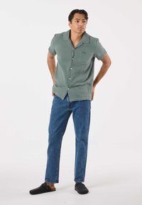 Camicia verde a maniche corte con bottoni e finitura testurizzata, abbinata a jeans blu a gamba dritta e zoccoli neri. Design semplice, senza motivi.