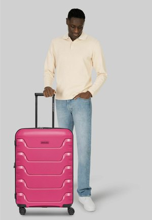Homme en chemise beige à manches longues et jean tenant la poignée d'une grande valise à roulettes rose sur fond gris.