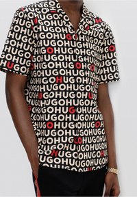 Camicia a maniche corte con un motivo nero, beige e rosso che presenta la parola "HUGO" ripetuta. Visibili i bottoni e i dettagli del colletto.