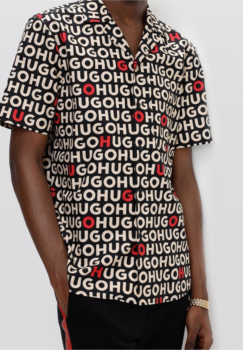 Camicia a maniche corte con un motivo nero, beige e rosso che presenta la parola "HUGO" ripetuta. Visibili i bottoni e i dettagli del colletto.
