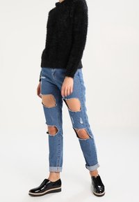 Schwarzer flauschiger Pullover, hellblaue zerrissene Jeans mit großen Löchern und schwarze Lackschuhe mit beigefarbener Plateausohle. Umgekrempelte Hosenbeine.
