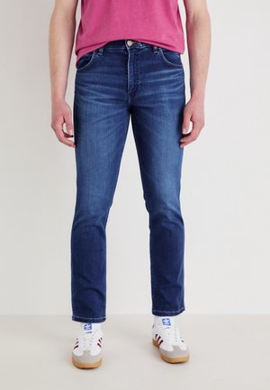 Personne portant un jean slim bleu foncé, des baskets blanches et rouges, et un t-shirt rose, debout devant un fond clair uni.