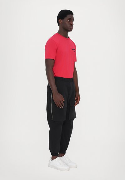 Red short-sleeve t-shirt with black text, paired with black cargo-style pants and white sneakers. Model stands in profile against a plain background.
