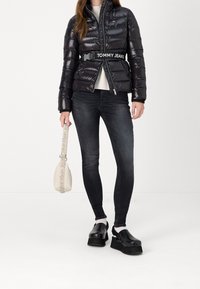 Chaqueta acolchada negra con acabado brillante y cierre de cremallera. Jeans oscuros de corte ajustado. Zapatos de plataforma negros. Pequeño bolso blanco con correa y acentos de logo.