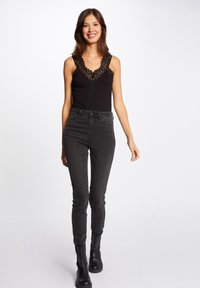 Body nero con inserti in pizzo, abbinato a jeans neri a vita alta e stivali neri alla caviglia. La modella posa su uno sfondo bianco.