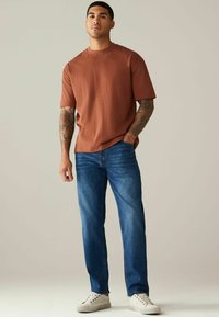Bruine T-shirt met korte mouwen, gecombineerd met blauwe denim jeans. Casual schoenen hebben een gestructureerde witte stof met een rubberen zool.