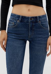 Jeans de mezclilla en azul oscuro, con una cintura de altura media, diseño clásico de cinco bolsillos y botón y remaches de tono cobre para un detalle adicional.