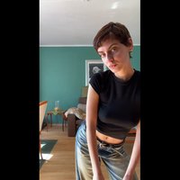 Fekete, testhez simuló póló, kék farmernadrág övvel, benti térben, égkék fal előtt állva. A természetes fény kiemeli a textúrákat és a színeket.