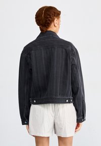 Giacca di jeans nera con strisce verticali, dettagli con bottoni e un design leggermente corto. Le maniche sono lunghe e aderenti.
