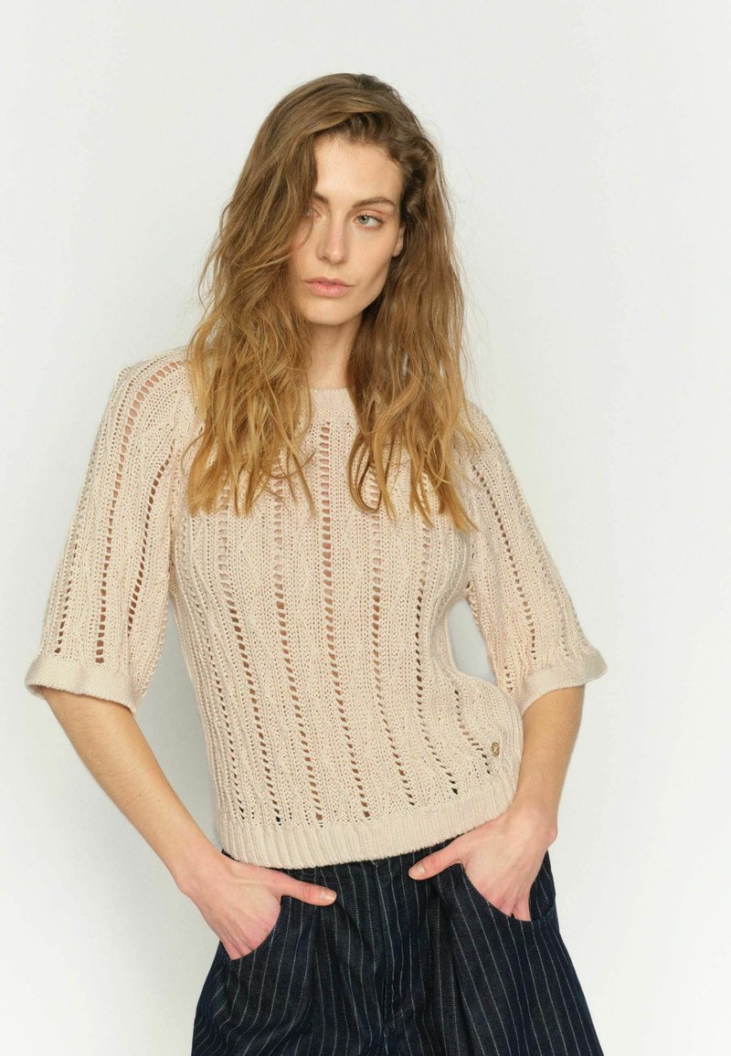 Femme aux cheveux ondulés portant un pull beige en maille à manches courtes et un pantalon foncé à fines rayures, debout les mains dans les poches contre un fond uni.