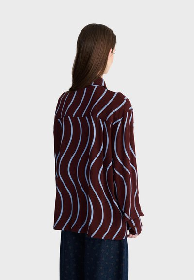 Personne aux longs cheveux bruns porte une chemise bordeaux ample avec des rayures ondulées verticales bleu clair, vue de dos sur un fond uni.