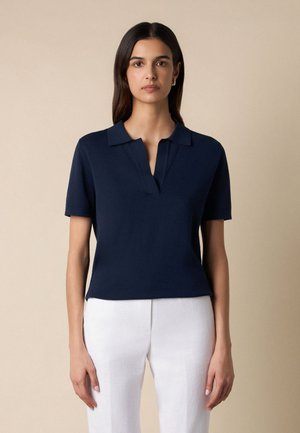 Kobieta o długich, ciemnych włosach, ubrana w granatową koszulkę polo z krótkim rękawem i białe spodnie, stojąca na tle beżowego tła.