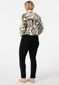Femme aux cheveux blonds courts se tenant de dos, portant un chemisier à manches longues à motifs de feuilles, un pantalon noir et des chaussures plates beiges ouvertes à l'arrière.