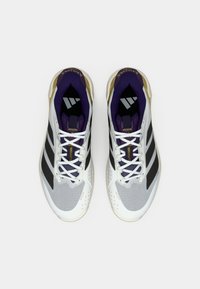 Białe buty sportowe z czarnymi paskami i fioletowymi akcentami. Posiadają siateczkową teksturę, okrągły czubek i płaskie sznurowadła. Złote detale w pobliżu podeszwy.