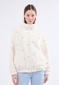 Puszysta kremowa kurtka z niebieskimi haftowanymi wzorami w kształcie kokardy, z wysokim kołnierzem, zamek z przodu i dwoma bocznymi kieszeniami. Miękka w dotyku, luźny krój.