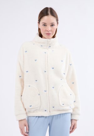 Chaqueta de crema difusa con patrones bordados de lazo azul, cuello alto, cierre frontal y dos bolsillos laterales. Textura suave, corte oversized.